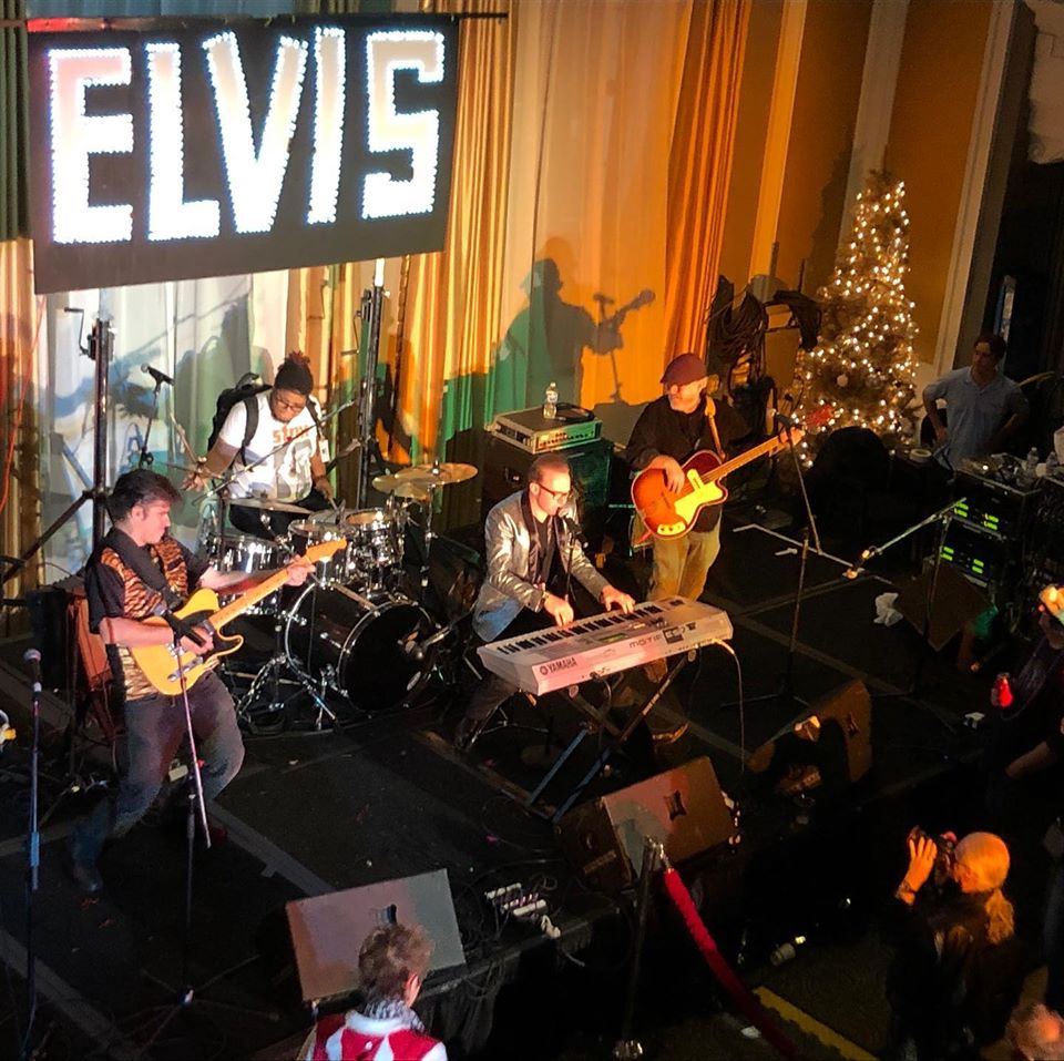 4 band members playing instruments on stage under ELVIS sign