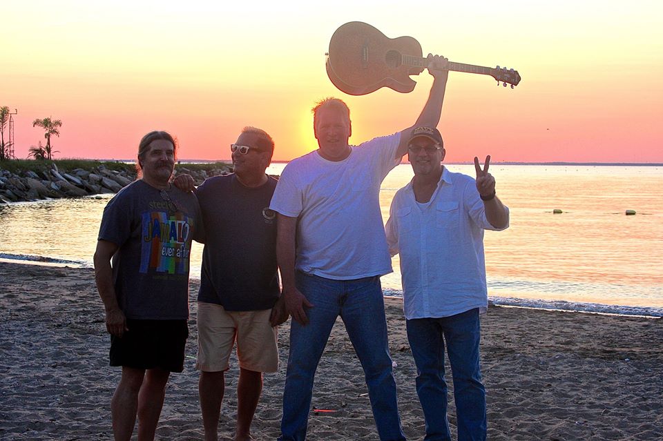 4 men standing on beach with one holding guitar above his head